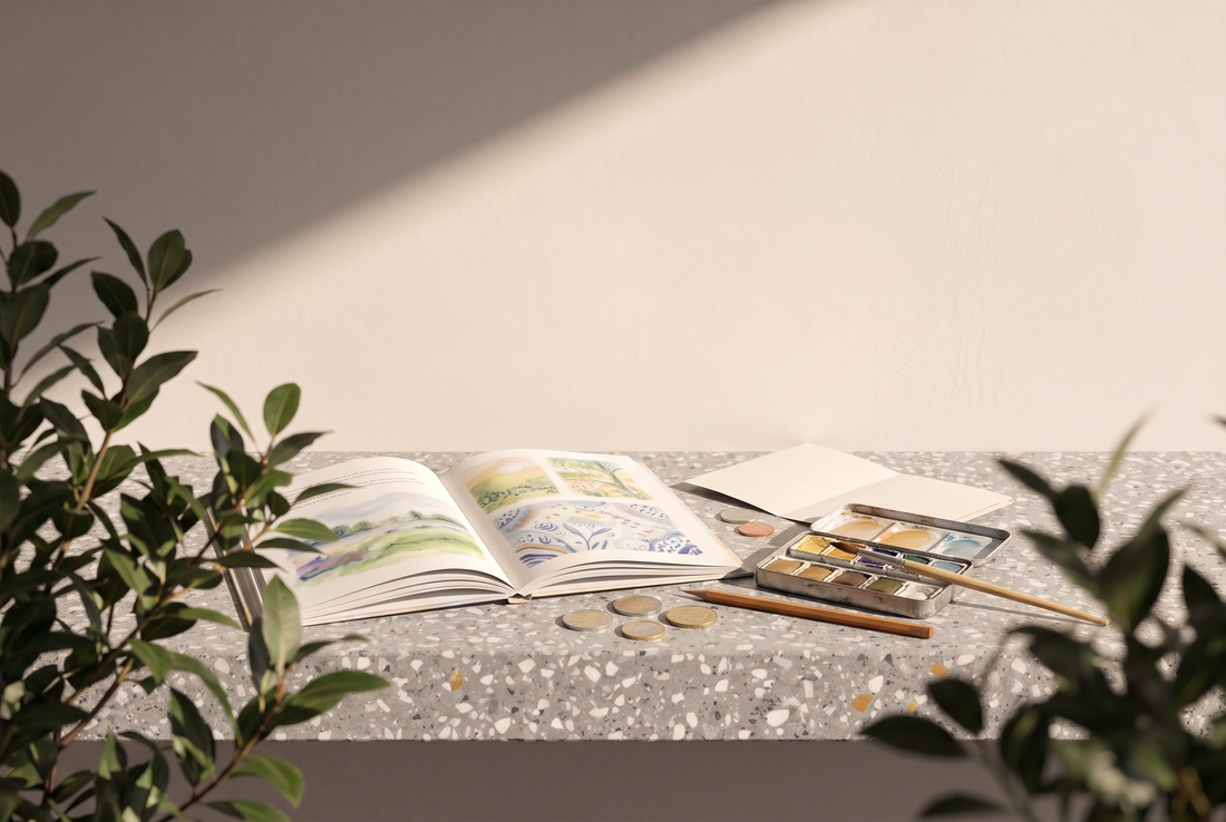 Aufgeschlagenes Bilderbuch mit Aquarellillustrationen, Aquarellkasten mit Pinsel, Bleistift, gefaltetes Blatt Papier und einige Euro-Münzen auf einer Terrazzokante vor heller Wand in der Sonne