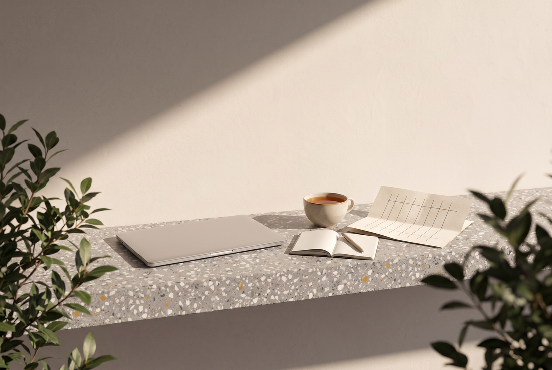 Zugeklappter Laptop, aufgeschlagenes Notizbuch mit Stift, Tasse Tee und gefalteter Wochenplan auf einer Terrazzokante vor heller Wand in der Sonne