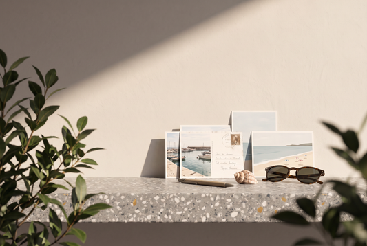Postkarten mit Hafen- und Strandmotiven, Kugelschreiber, Muschel und Sonnenbrille auf einer Terrazzokante vor heller Wand in der Sonne