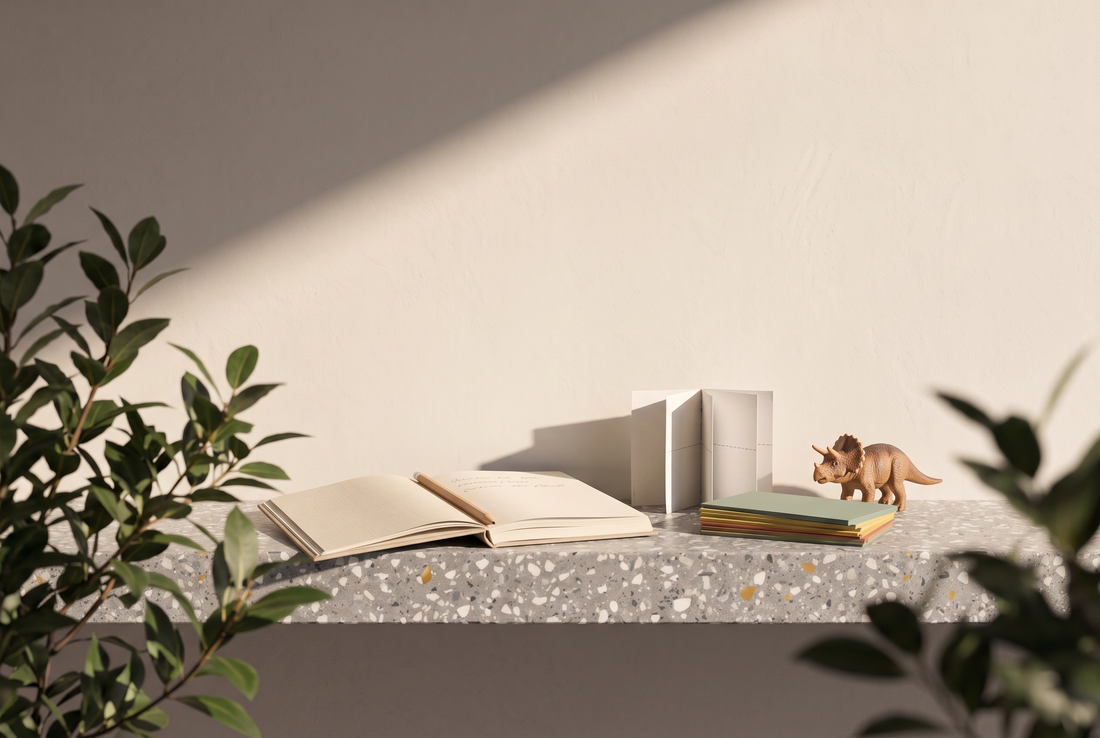 Aufgeschlagenes Notizbuch mit Bleistift, ein gefaltetes Papier-Probebüchlein, ein Stapel bunter Karteikarten und ein kleiner Spielzeugdinosaurier auf einer Terrazzokante vor heller Wand in der Sonne