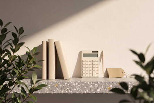 Taschenbücher mit schlichten Umschlägen, ein heller Taschenrechner, ein Bleistift und ein leeres Preisschild aus Kraftpapier auf einer Terrazzokante vor heller Wand in der Sonne