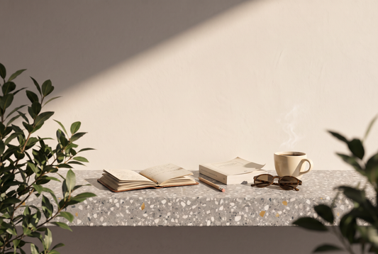 Aufgeschlagenes Notizbuch, Bleistift, Taschenbuch, Sonnenbrille und eine Tasse Kaffee auf einer Terrazzokante vor heller Wand in der Sonne
