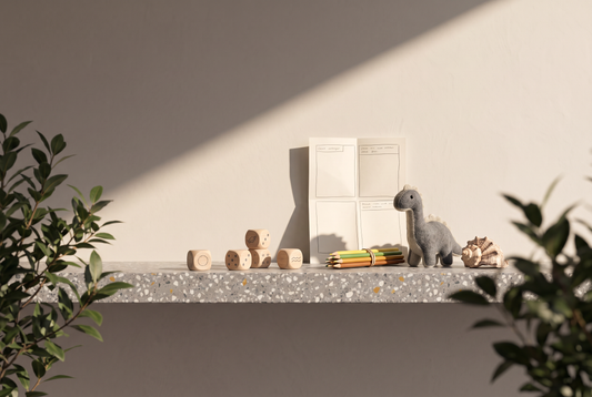 Holzwürfel mit gezeichneten Symbolen, ein Blatt mit leeren Comicfeldern, Buntstifte, ein Plüschdinosaurier und eine Muschel auf einer Terrazzokante vor heller Wand in der Sonne