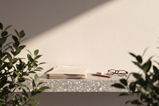 Stapel Manuskriptseiten, roter Bleistift, Radiergummi und Lesebrille auf einer Terrazzokante vor heller Wand in der Sonne