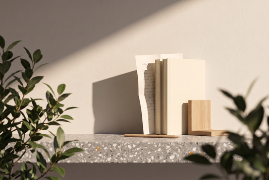Taschenbücher mit schlichten Umschlägen, ein gefaltetes Blatt, ein Bleistift und eine hölzerne Buchstütze auf einer Terrazzokante vor heller Wand in der Sonne