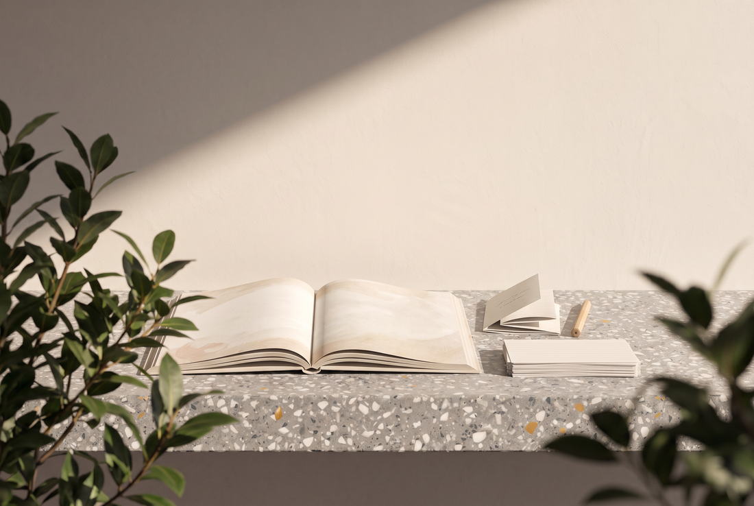 Aufgeschlagenes Bilderbuch mit zarten Aquarellseiten, ein gefaltetes Papierbüchlein, ein Bleistift und ein Stapel Karteikarten auf einer Terrazzokante vor heller Wand in der Sonne