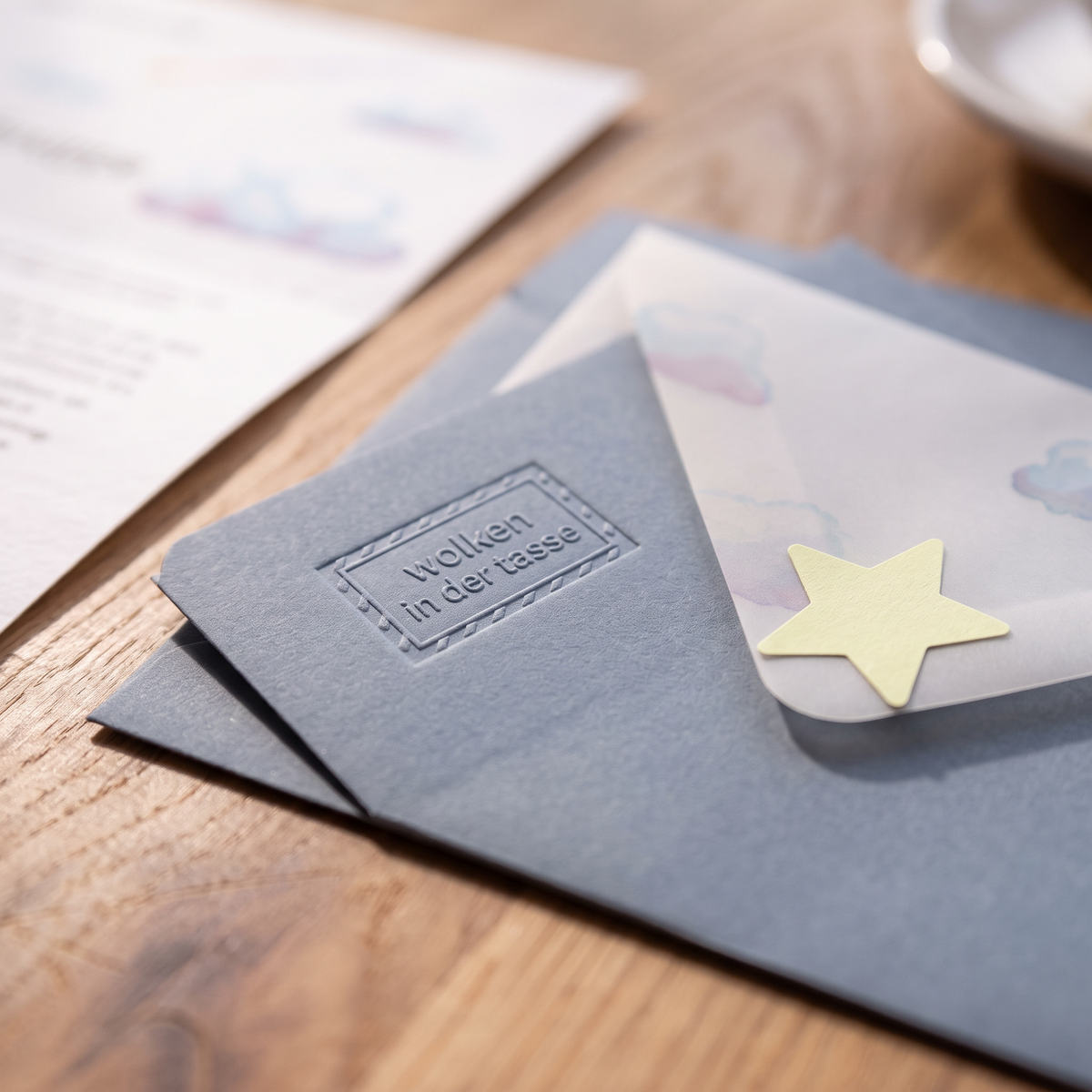 Graublaue Umschläge mit Blindprägung „wolken in der tasse“ und Transparentumschlag mit Stern auf Eichenholz, schräg fotografiert