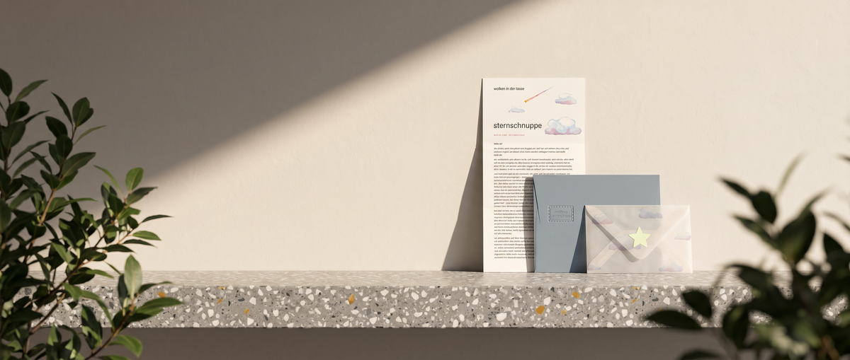 Briefabo wolken in der tasse: Brief „sternschnuppe“, graublauer Umschlag mit Prägung und Transparentumschlag mit Stern auf einer Terrazzokante vor warmweißer Wand in der Abendsonne, vorn unscharfes Grün